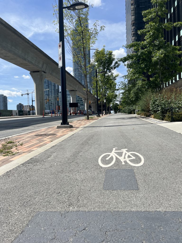 A protected bike lane is nestled between residences and a road with a SkyTrain elevated guideway running alongside it.